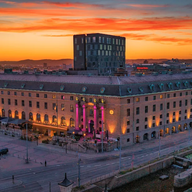 Ett stort hotell i klassisk stil upplyst i rosa och gult under en färgstark solnedgång i staden.