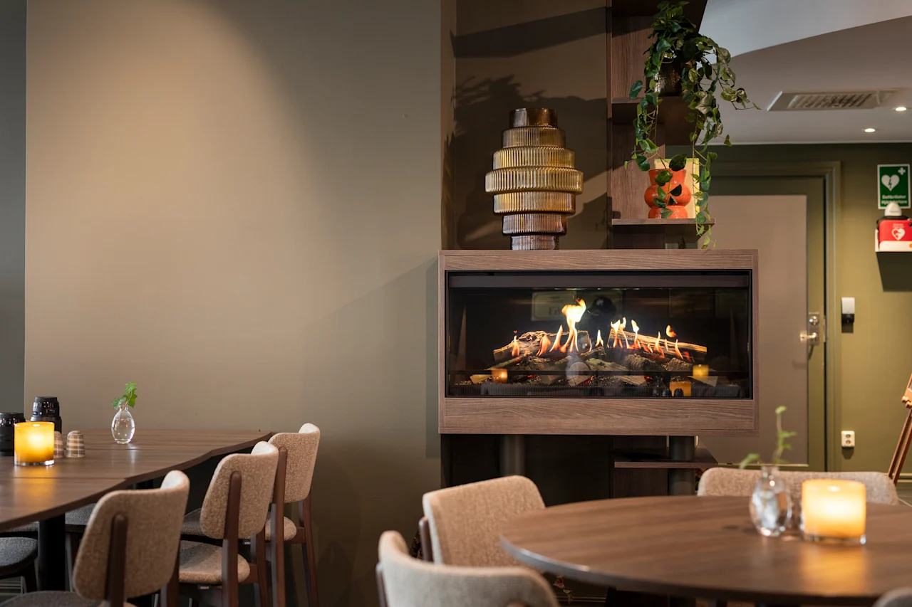 Viihtyisä ravintola- tai lounge-alue, jossa on moderni takka, pöytiä, tuoleja ja sytytettyjä kynttilöitä, luoden lämpimän ja kutsuvan tunnelman.
