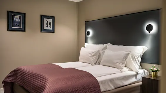 A neatly made bed with white sheets and a purple blanket stands against a taupe wall, illuminated by two wall sconces. Framed penguin art decorates the wall.