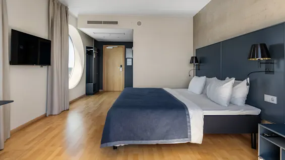 Et moderne hotelværelse med en stor seng, trægulv, et vægmonteret TV og et unikt rundt vindue med gardiner.