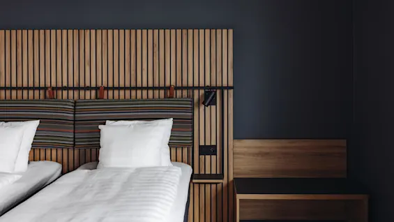 Two neatly made twin beds with white linens stand against a modern wood-panel headboard in a minimalist room, featuring a dark wall and a small wooden nightstand.