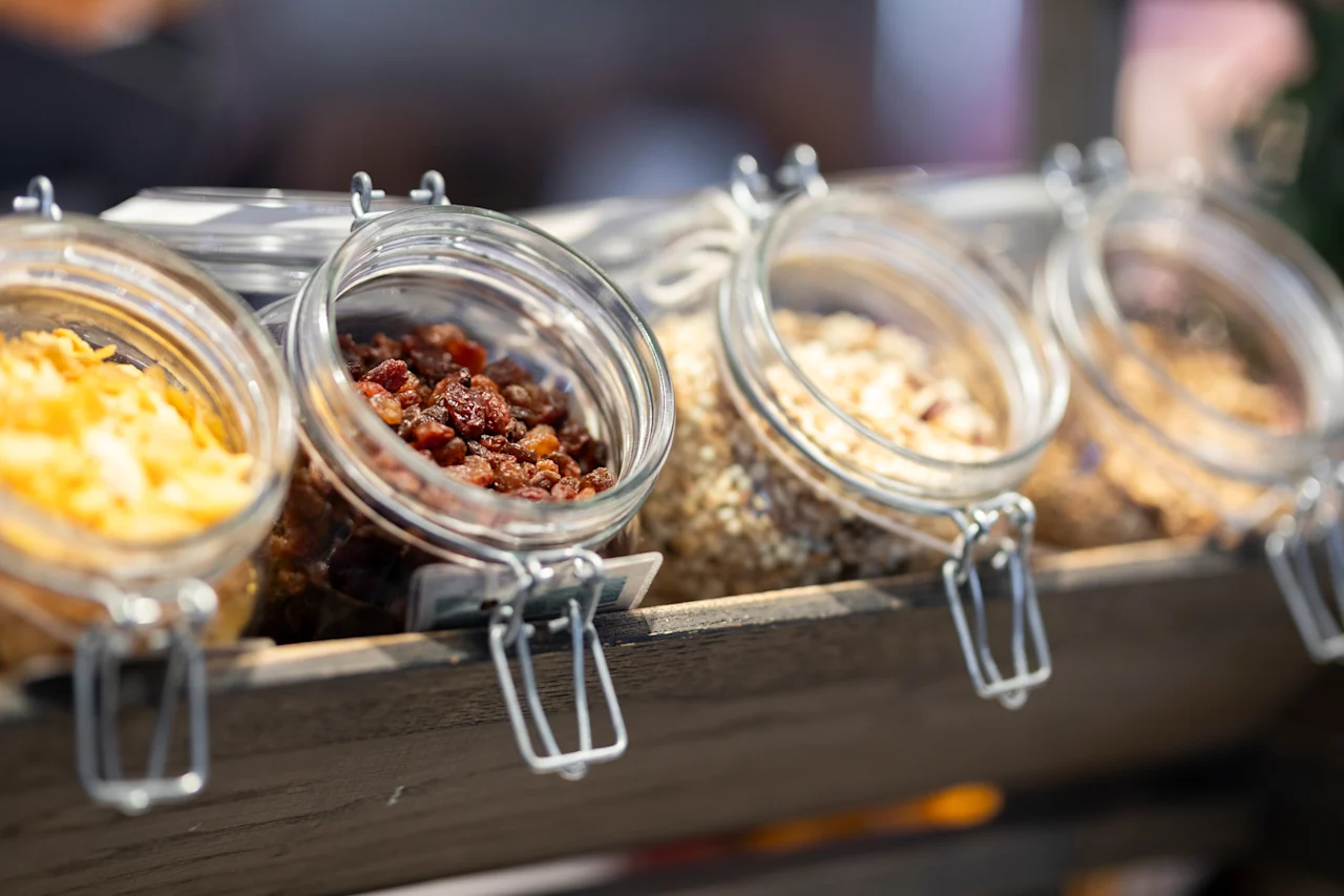 Lähikuva useista lasipurkeista, jotka on täytetty erilaisilla aamiaistarvikkeilla, kuten kuivatuilla hedelmillä ja muroilla, esillä puutelineellä.