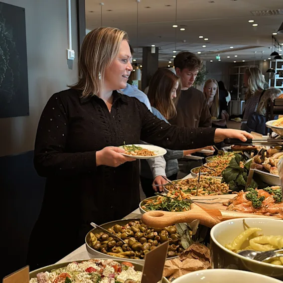 En person forsyner seg fra en rikholdig buffet med ulike salater og retter, omgitt av andre gjester.