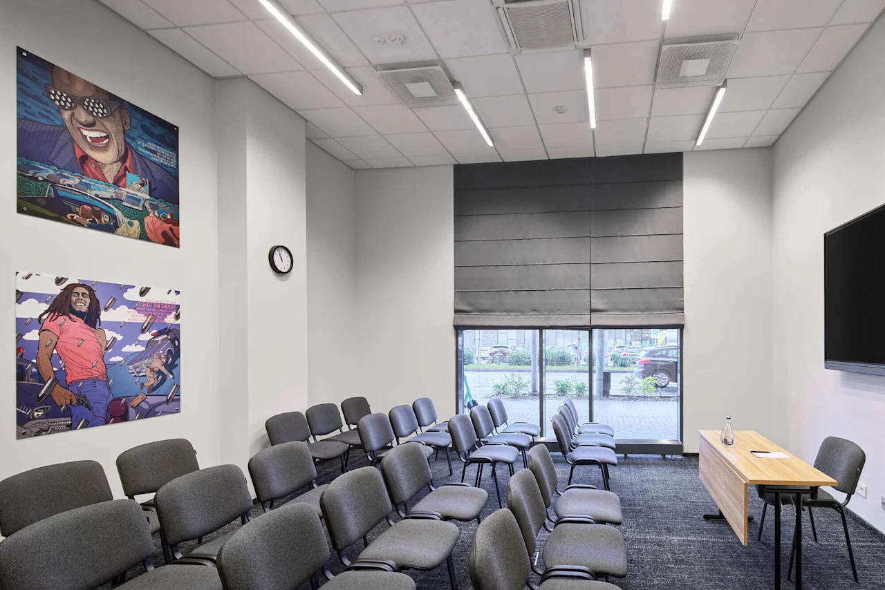 Rows of gray chairs face a wooden desk and a wall-mounted screen in a conference room. Art posters and a wall clock adorn the walls, with light streaming through large windows.