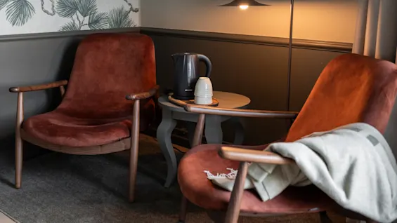 Kaksi viihtyisää nojatuolia ja pieni pöytä, jolla on vedenkeitin ja kupit, valaistuna lämpimällä lampulla, luoden kutsuvan tilan.
