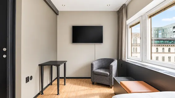 Bright hotel room featuring a desk, armchair, and wall-mounted TV next to a window with a city view.