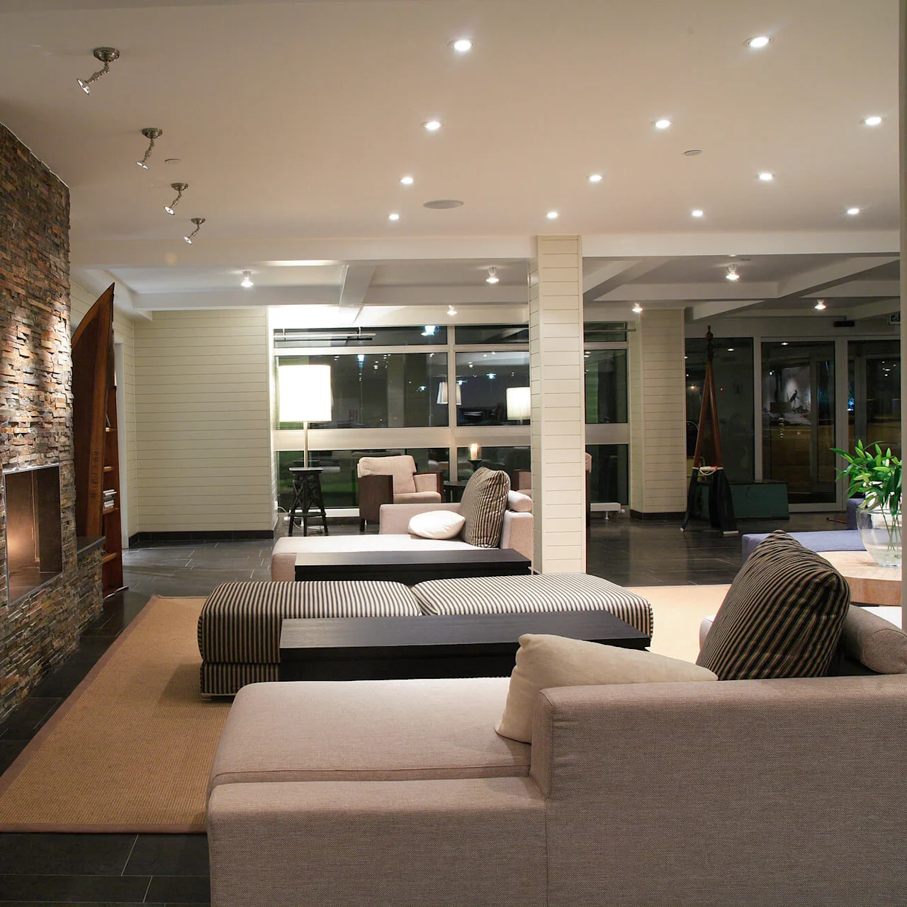 Modern, cozy living room features a stone fireplace, adjacent couch seating, soft lighting from ceiling spotlights and lamps, with a large window providing night views. Wall art depicts a sailboat.