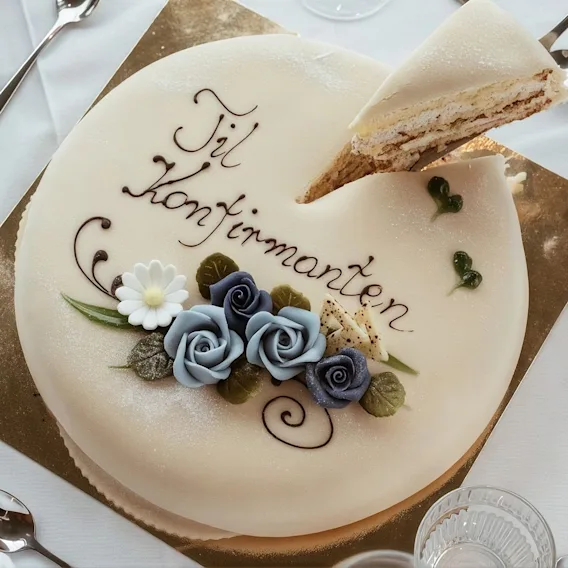 Servering av et stykke hvit Til Konfirmanten kake pyntet med bla roser pa et pent dekket bord