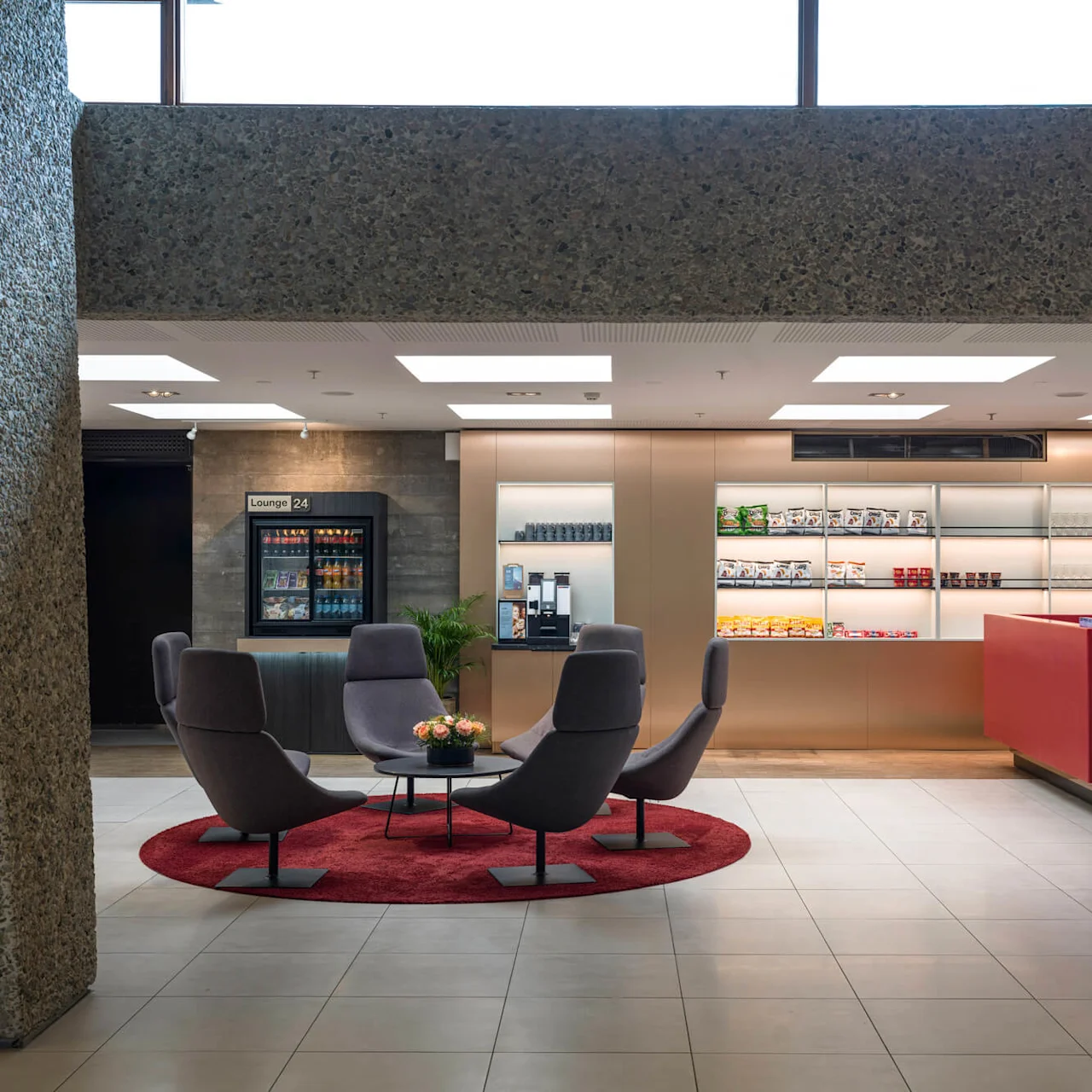 Seating area with five gray chairs surrounds a small table with flowers, situated on a red rug. Nearby, snack shelves and a sign reading "Lounge 24" provide a relaxing context.