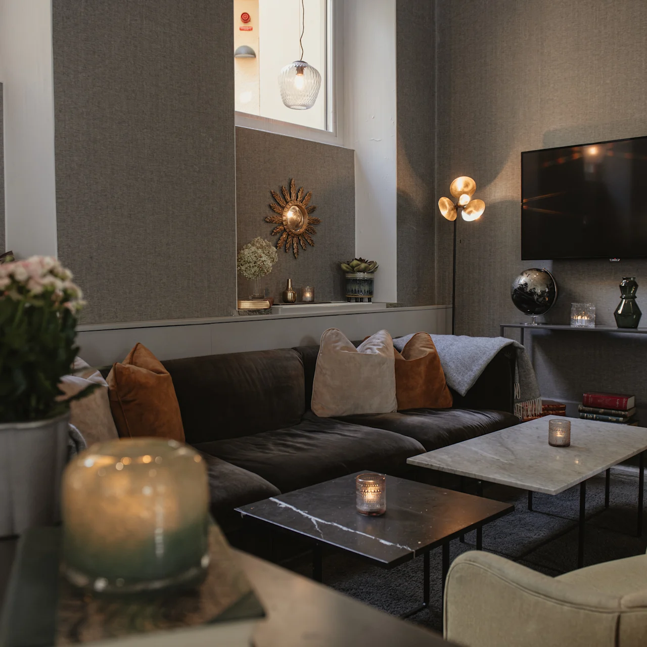 Ett mysigt, modernt vardagsrum har en mörk soffa med kuddar, en väggmonterad TV, dekorativa lampor och en blomvas på ett bord, vilket skapar en varm atmosfär.