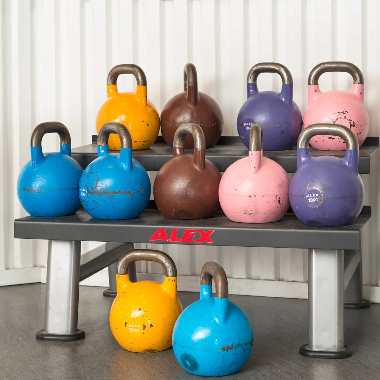 Colorful kettlebells resting on a gym rack against a white corrugated metal wall. ALEX and various weights marked on them, including 18KG.