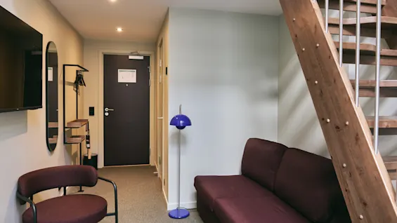 A modern living space with a dark red sofa, a blue floor lamp, and a wooden staircase leading to an upper level.