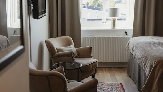 Viihtyisä hotellihuone, jossa on kaksi mukavaa nojatuolia, sänky ja ikkuna, josta tulee luonnonvaloa, luoden rentouttavan tunnelman.