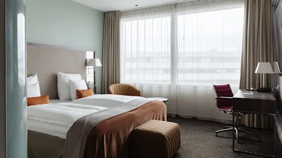 Moderni hotellihuone, jossa mukava sänky, työpöytä tuolineen ja suuret ikkunat ohuilla ja pimennysverhoilla.