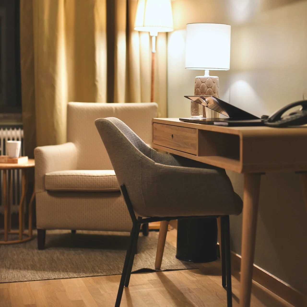 En grå stol står vid ett skrivbord av trä med en vit lampa, i ett varmt upplyst rum med beige gardiner, en fåtölj och trägolv.