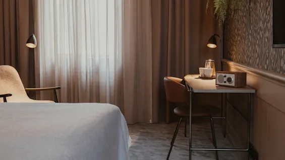 Lämmin ja kutsuva hotellihuone, jossa on mukava sänky, työpöytä radiolla ja pehmeä luonnonvalo.