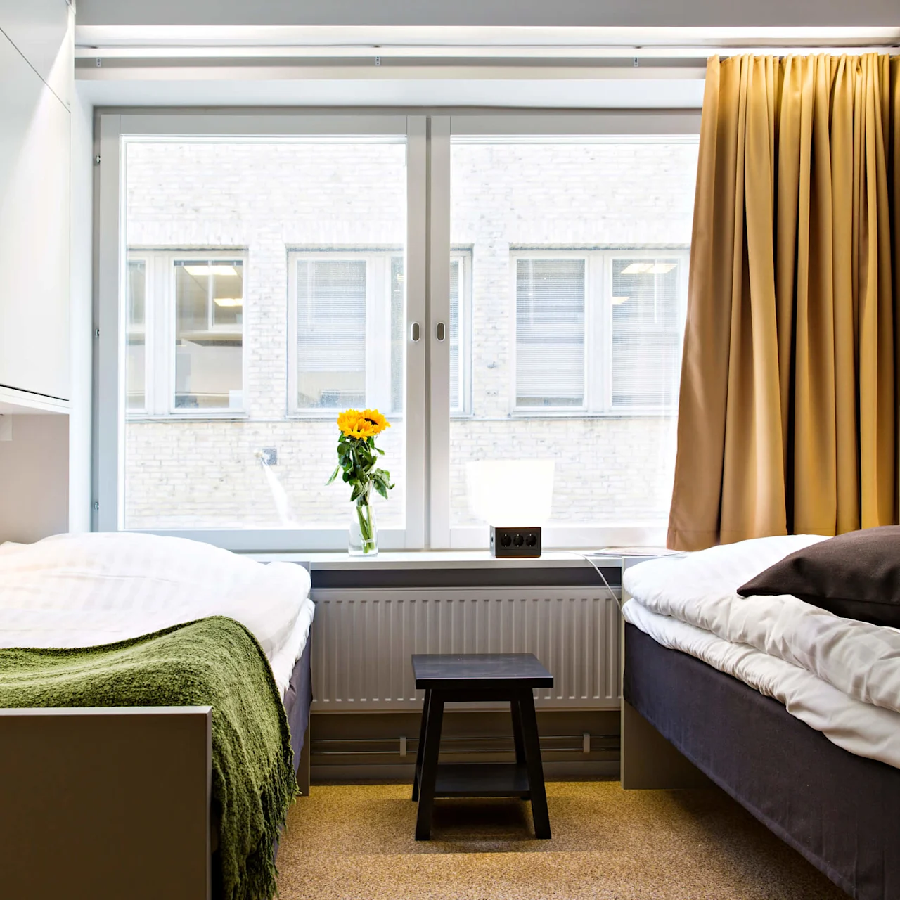 To enkeltsenger står i et rom med et stort vindu. En gul blomst i en vase står på vinduskarmen, med beige gardiner som rammer inn scenen.