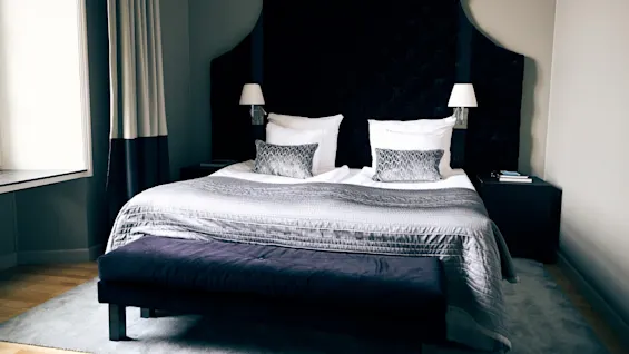 A bed with a dark tufted headboard and gray bedding stands in a minimalist room. Two white lamps are mounted on either side. A bench and bedside tables complete the setting.