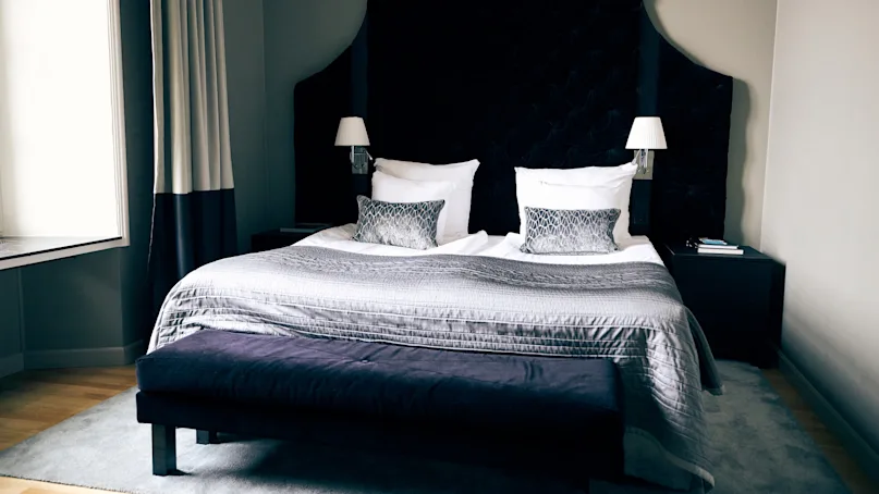 A bed with a dark tufted headboard and gray bedding stands in a minimalist room. Two white lamps are mounted on either side. A bench and bedside tables complete the setting.