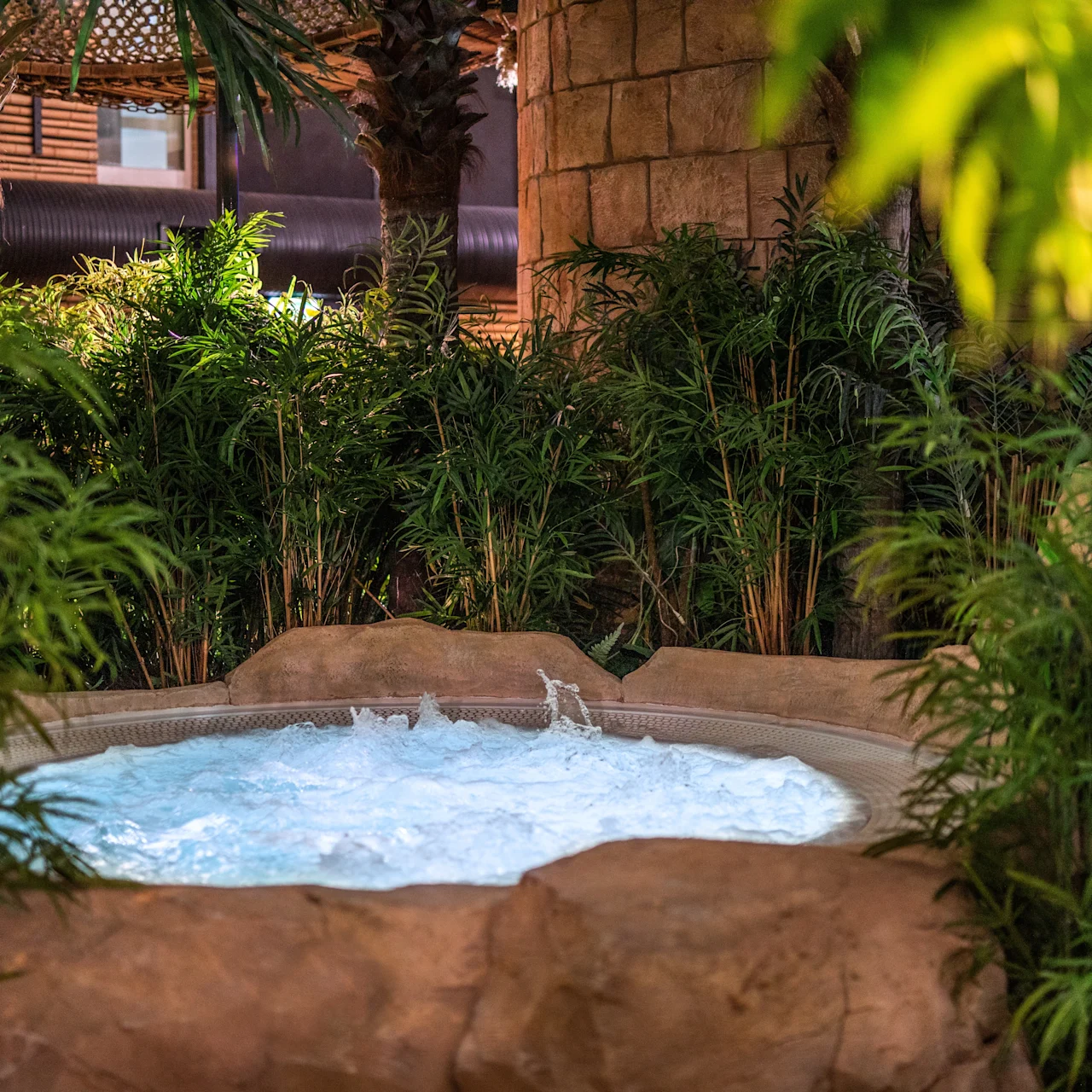 A bubbling hot tub surrounded by lush green plants creates a tropical oasis atmosphere, set against a backdrop of wooden structures and stone walls.