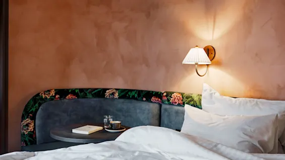A neatly made bed with white sheets is beside a wall-mounted lamp and nightstand holding a book and a cup. The backdrop is a textured peach-colored wall.