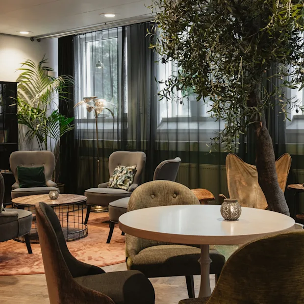 A comfortable and inviting lounge area at Home Hotel Uppsala with various seating, tables, and lush green plants.