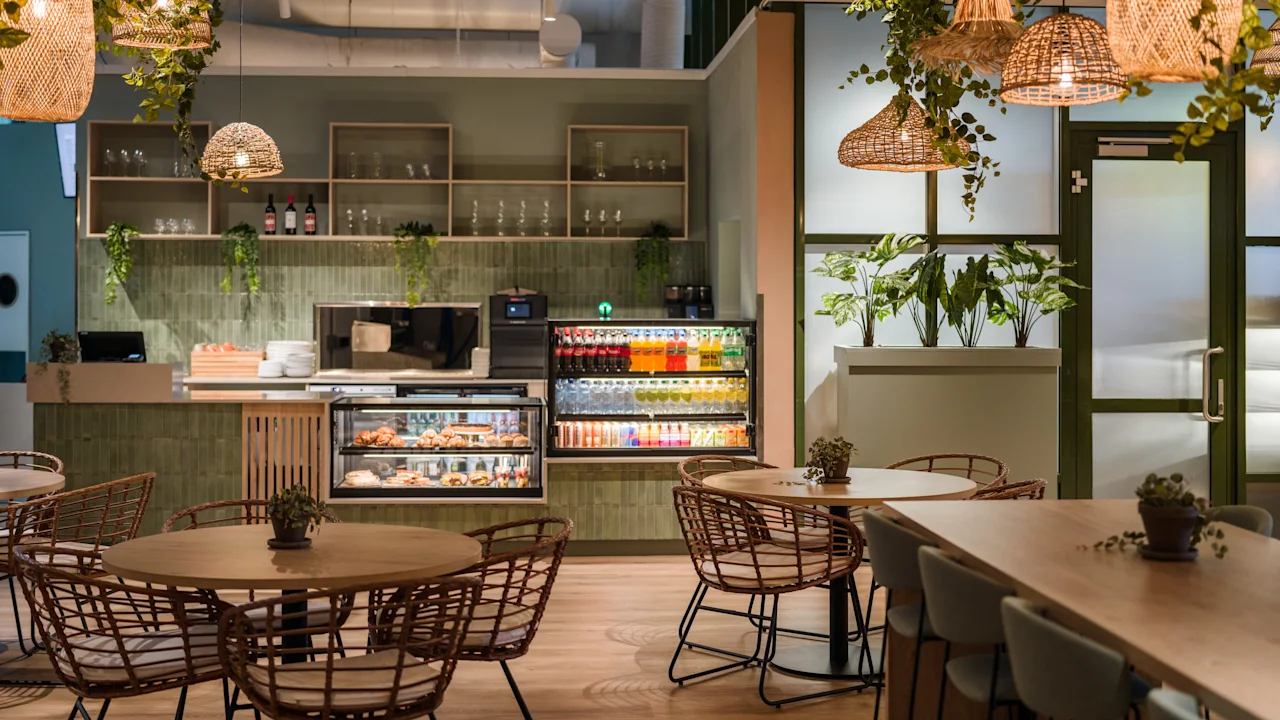 Bord og stoler står under hengende planter og kurvlamper. En disk viser snacks og drikkevarer. Hyller holder glass og flasker i denne moderne kaféinnstillingen.