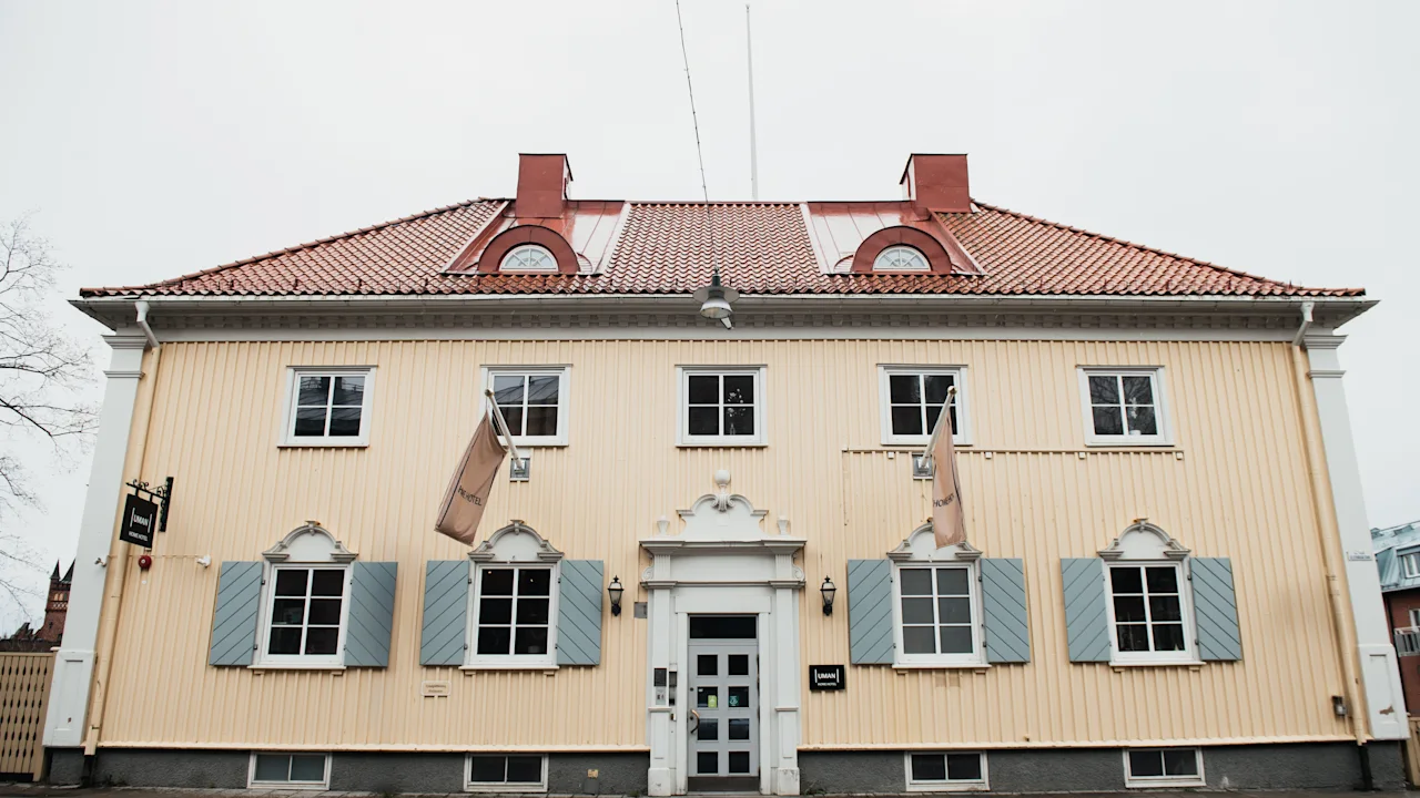 A beige wooden building with a red tiled roof, features six windows with blue shutters and three flags, standing on a paved street. 
