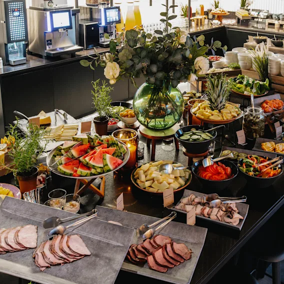 Buffet table displays assorted foods including sliced meats, fruits, and vegetables, surrounded by decorative plants. Two drink dispensers are visible in the background with stools placed around the table.