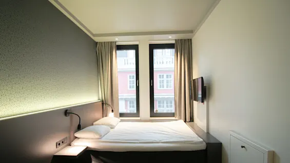 A neatly made bed sits in a small, minimalist room with a wall-mounted TV. Two large windows with curtains let in natural light.