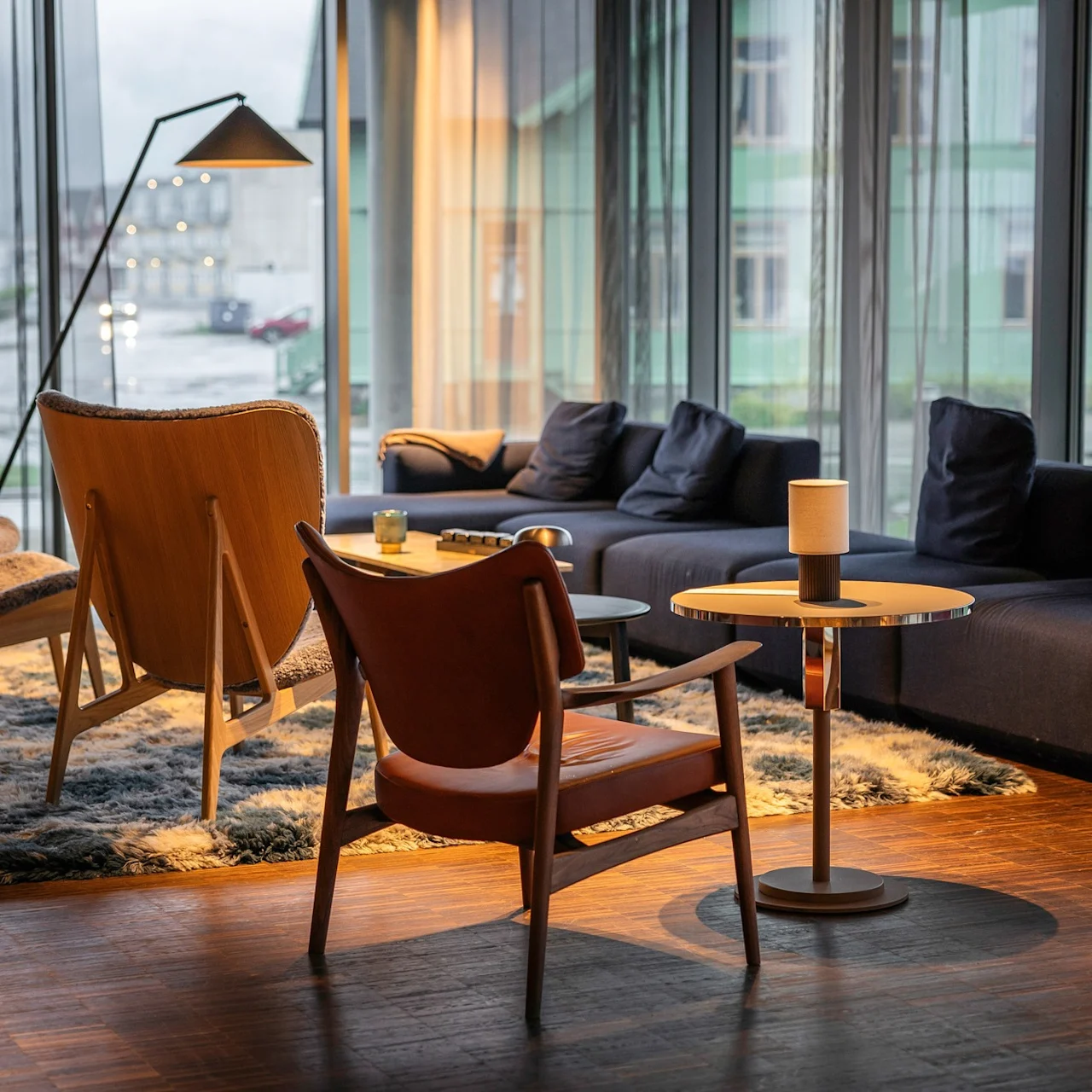 En hyggelig lounge har moderne lænestole omkring små borde under varmt lys. Området ligger på et mønstret tæppe med udsigt over en regnfuld gade gennem store vinduer.