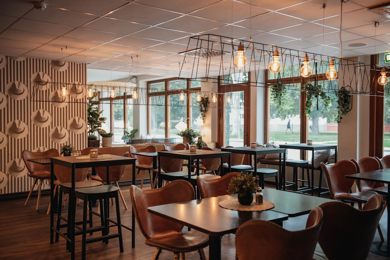 A cozy restaurant or cafe interior with a mix of high and low tables, warm lighting, and large windows overlooking greenery.