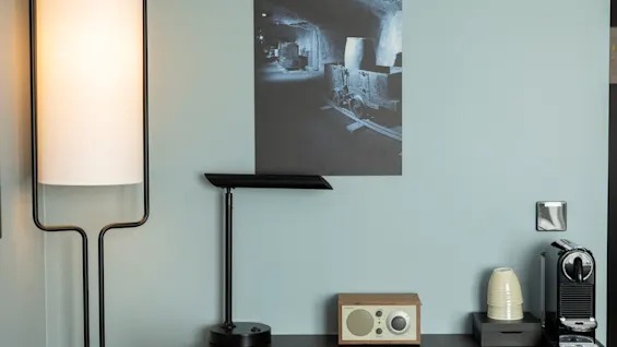 En koselig skrivebordsplass med moderne lampe, vintageradio og kaffemaskin, mot en lyseblå vegg med et historisk gruvefoto.