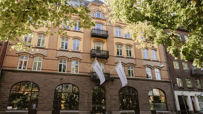 Home Hotel Norre Park in Halmstad, a charming building with flags, framed by green trees on a sunny day.