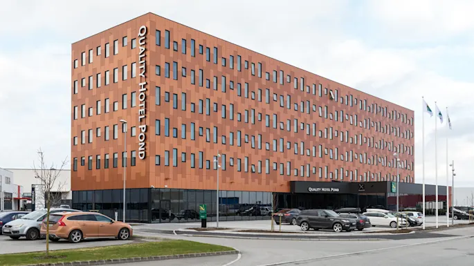 A modern, geometric hotel building stands with "QUALITY HOTEL POND" text in white. Cars are parked outside, and three flagpoles are visible. The sky is cloudy.