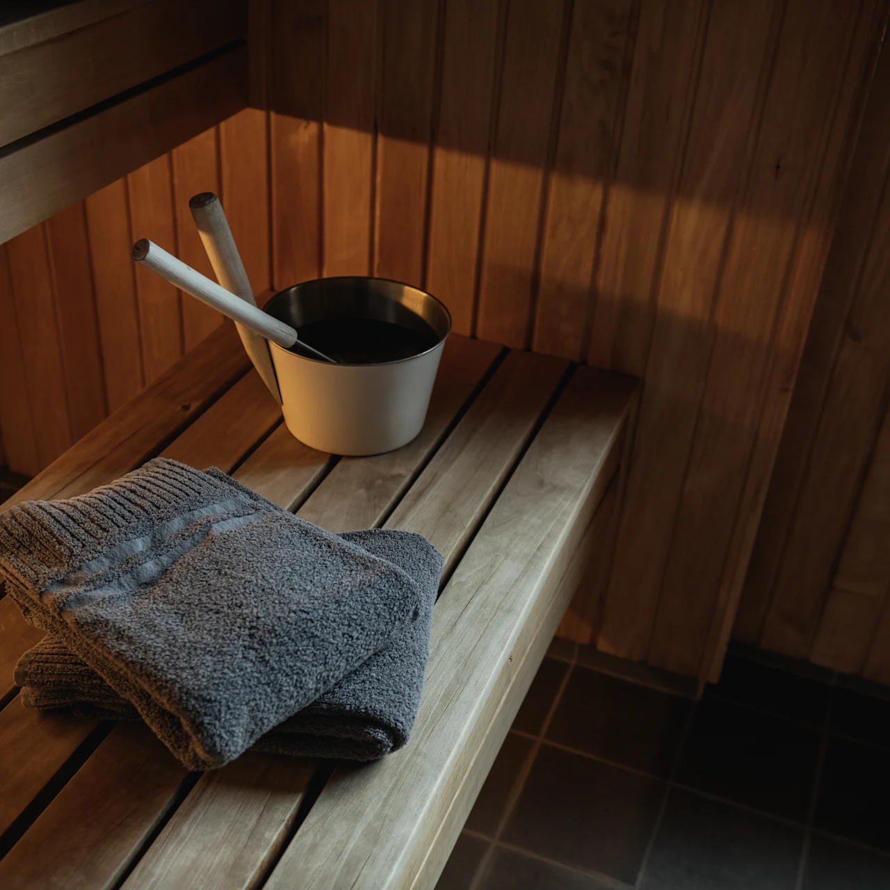 Towels lie folded on a wooden bench beside a white bucket with ladles in a sauna room with wooden walls and tiled floor.