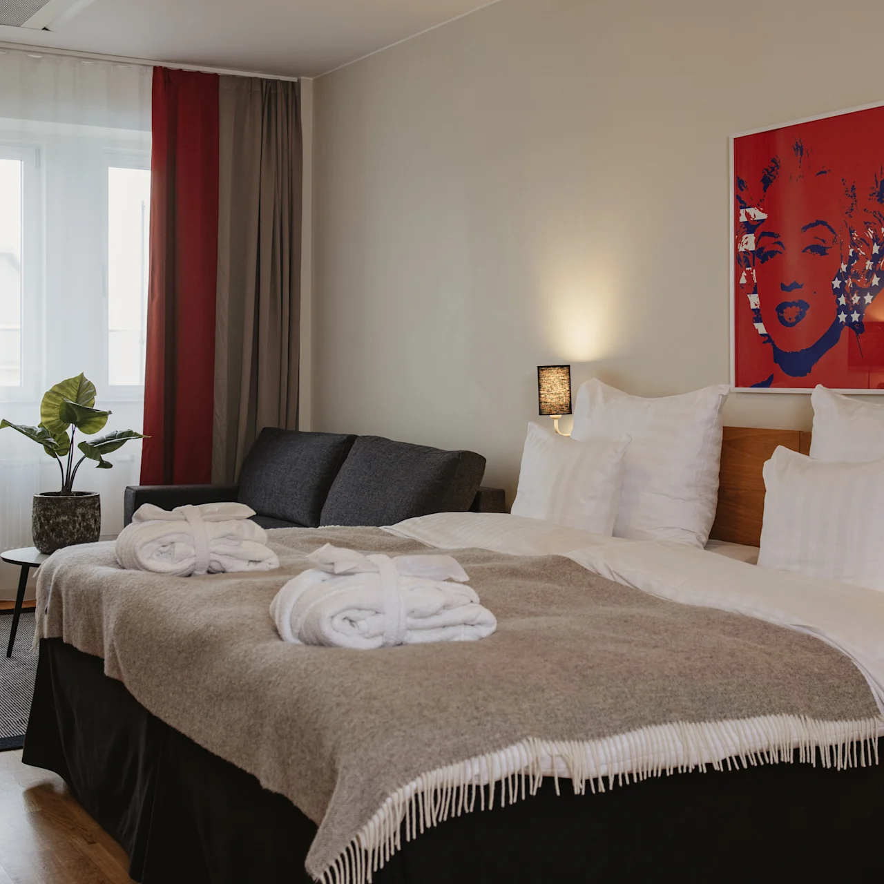 A neatly made bed with two robes lies in a cozy hotel room. A small couch and table with a plant are by the window. A colorful portrait adorns the wall.