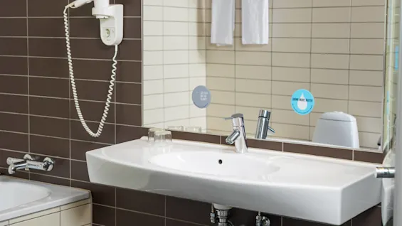 A white sink sits beneath a wall-mounted hairdryer in a tiled bathroom. The mirror displays text: "GO EASY ON THE PLANET" and "DRINK MORE WATER, generate less waste."