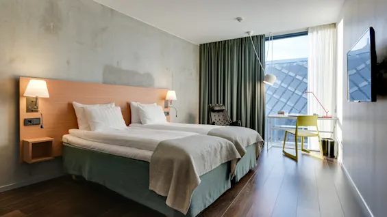 En pent og oppredd tvillingsover ligger i et moderne hotellrom, opplyst av vegglamper. I nærheten står et skrivebord og stol ved et vindu med forhengte gardiner.