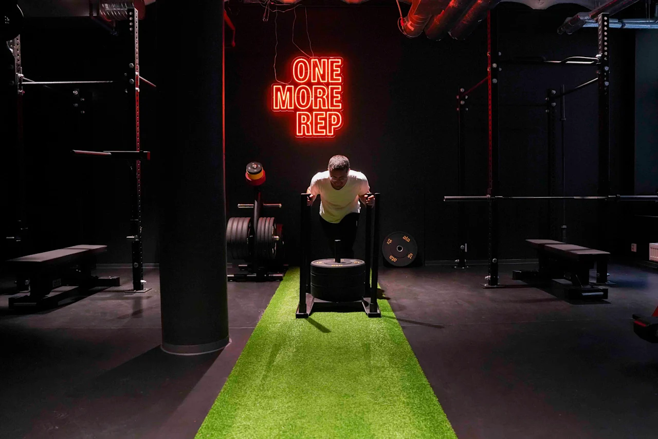 Mand skubber vægtet slæde på grønt græstæppe i fitnesscenter. "ONE MORE REP" i neonlys på sort væg. Omgivende udstyr inkluderer bænke og vægte.