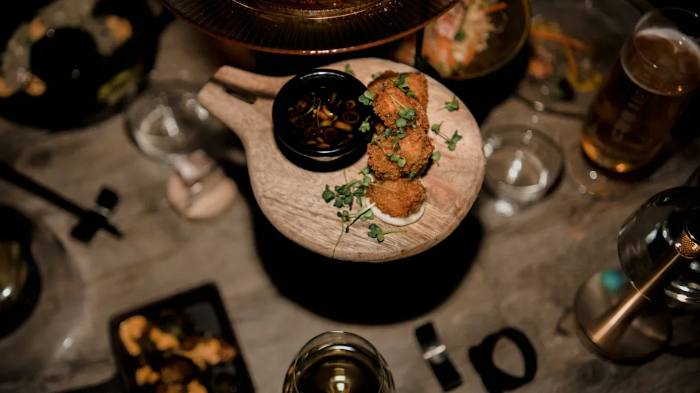 Brødkugler ligger på en træbakke, garneret med urter, ledsaget af en lille skål med dyppe sauce. Omgivelserne inkluderer forskellige retter, glas og bestik på et træbord.