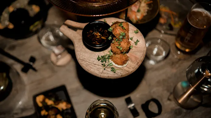 Brødkugler ligger på en træbakke, garneret med urter, ledsaget af en lille skål med dyppe sauce. Omgivelserne inkluderer forskellige retter, glas og bestik på et træbord.