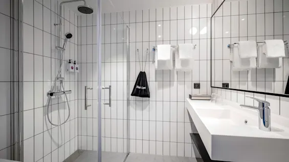 Ett modernt badrum har en duschkabin i glas med krombeslag, vita kaklade väggar och en bänk med ett handfat. Handdukar hänger i närheten, tillsammans med en svart påse märkt "Hårtork."
