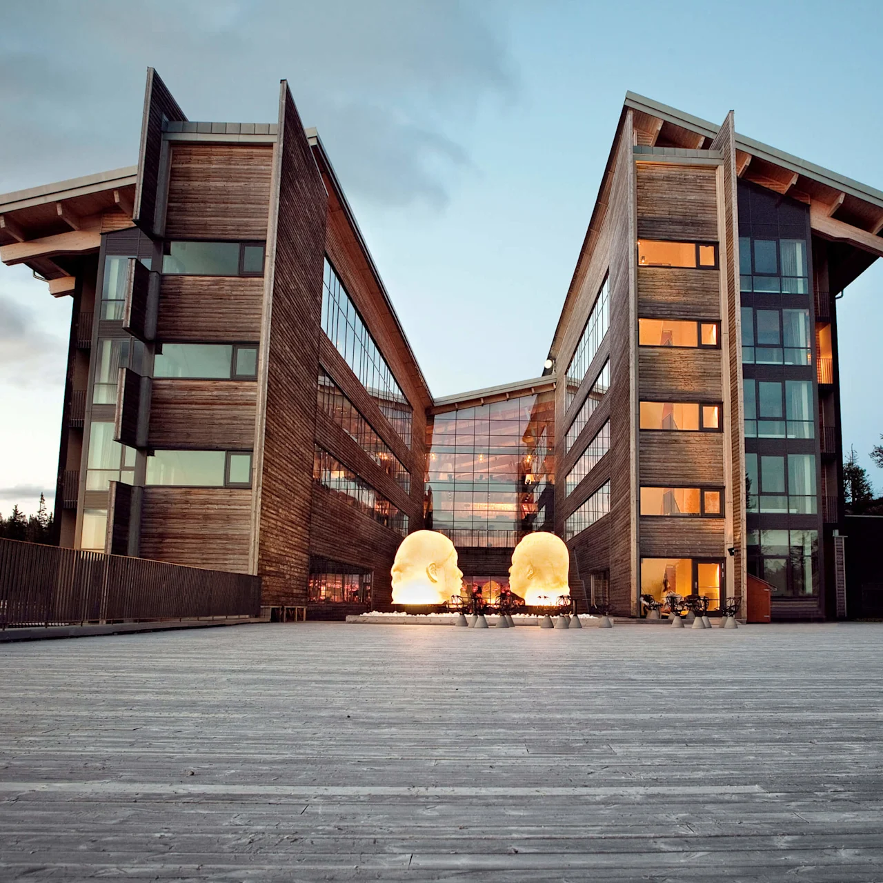 Modern building with symmetrical wings, illuminated large head sculptures in front, against a forested twilit sky.