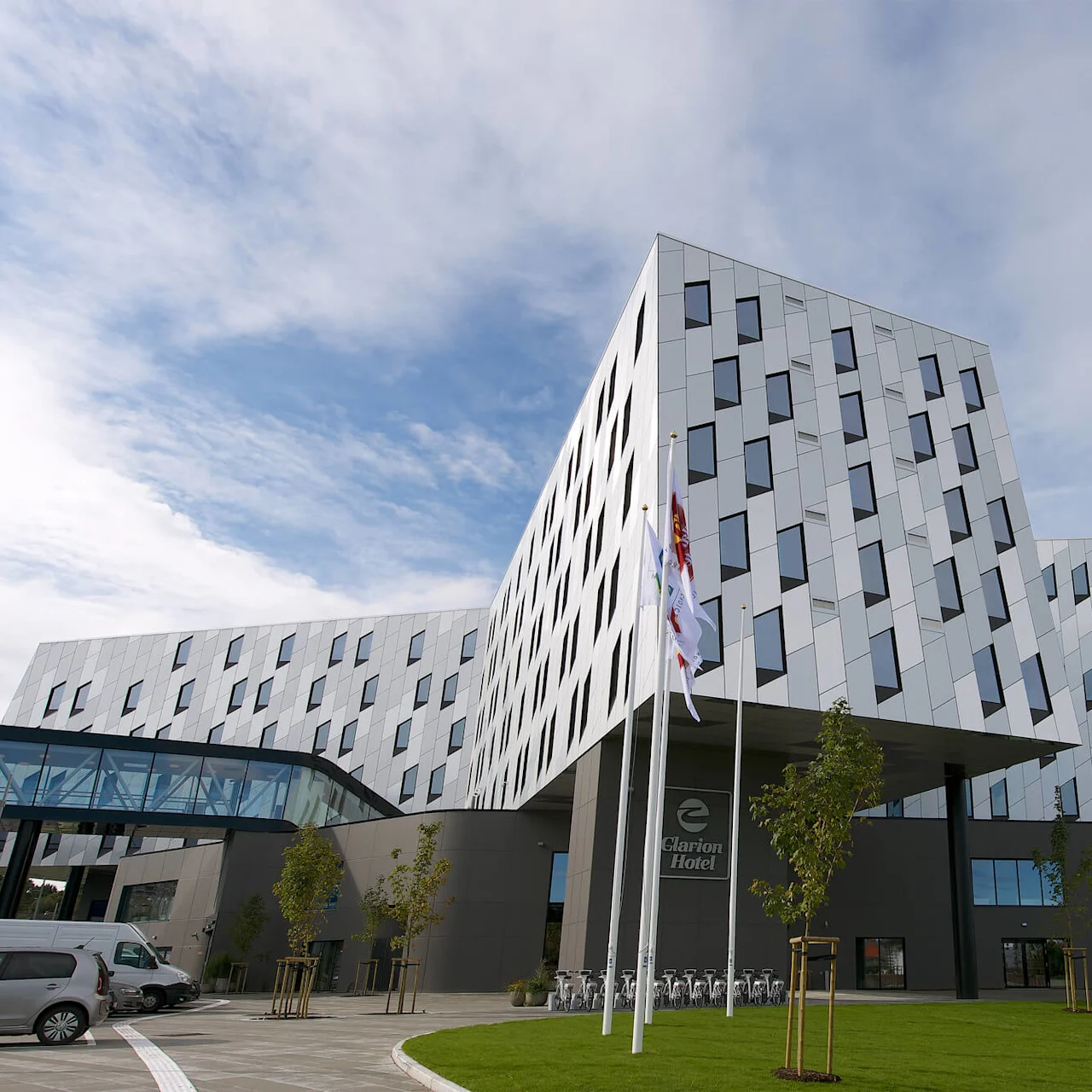 En modern, geometriskt utformad byggnad med vinklade väggar och många fönster står under en blå himmel. Vid ingången står "Clarion Hotel" och flera flaggor vajar i ett trädgårdsområde.
