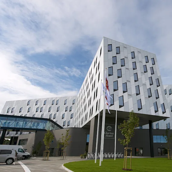 En moderne, geometrisk utformet bygning med skrå vegger og mange vinduer står under en blå himmel. Ved inngangen står det "Clarion Hotel" og flere flagg vaier i et hageområde.