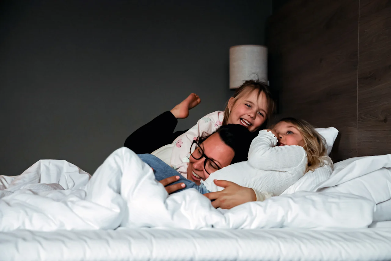 A person lies in bed with two children playfully embracing them, all smiling. The bed is covered with white sheets in a cozy bedroom setting.