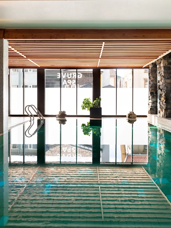 Innbydende innendørsbasseng på Quality Hotel Grand, Kongsberg, med naturlig lys, steinvegger, koselig sittegruppe og Grand Spa-refleksjoner.
