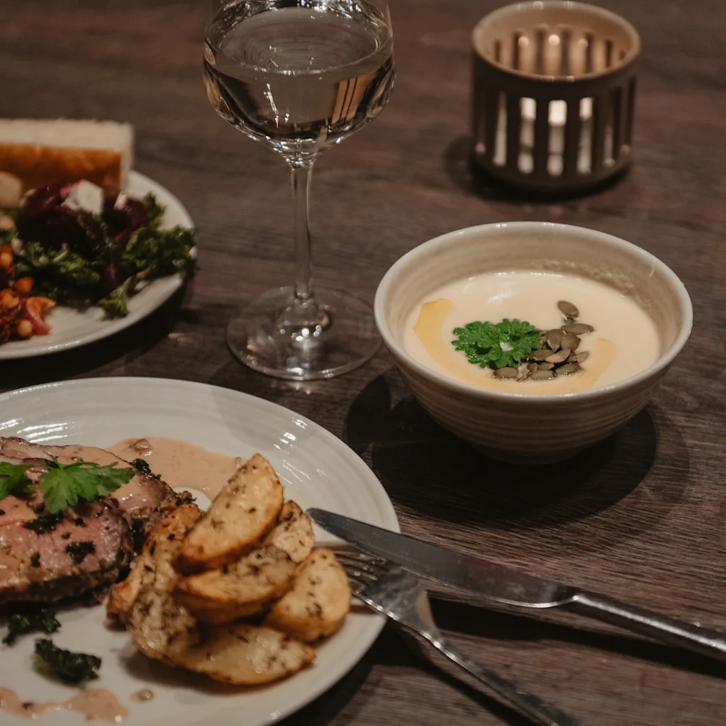 A cozy dinner setting with a main course of meat and potatoes, a creamy soup, a glass of white wine, and a lit candle.
