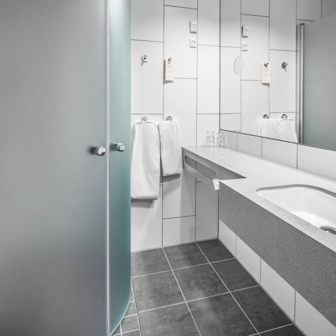 Modern bathroom featuring a frosted glass shower, a countertop with an integrated sink, and mirrored walls. White towels and a dark tiled floor complete the sleek design.